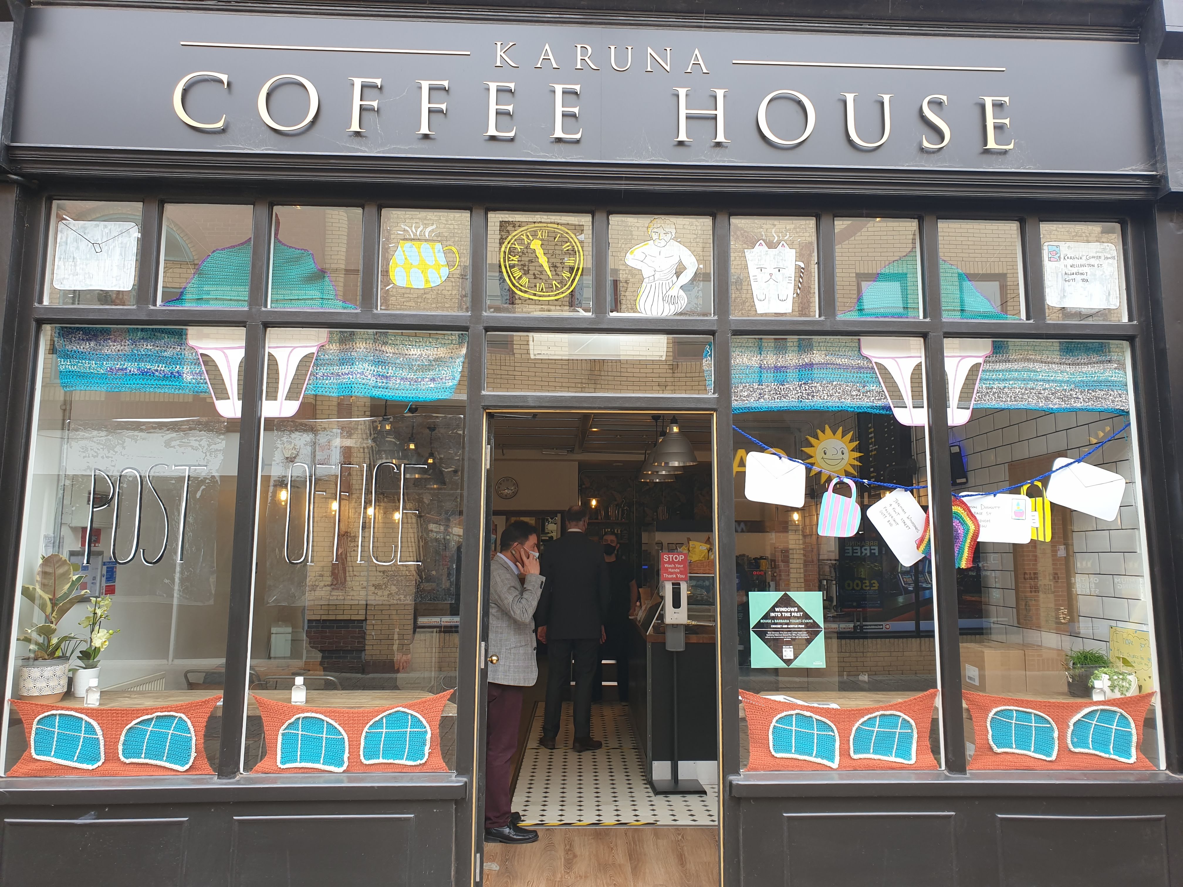 the front of a coffee shop. in the windows there are a number of decorations made of crochet and cartoon drawings on paper and clear plastic. along the top, there are two envelopes, two mugs, a clock, and a statue. a sign reading post office hangs in the left window.